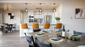 A dining area with a wooden table set for six, surrounded by chairs. A kitchen with modern appliances and pendant lighting is visible in the background. - Gorgeous Retreat with Mountain Views, Open Living, Game Room, Hot Tub (Glenwood Springs)