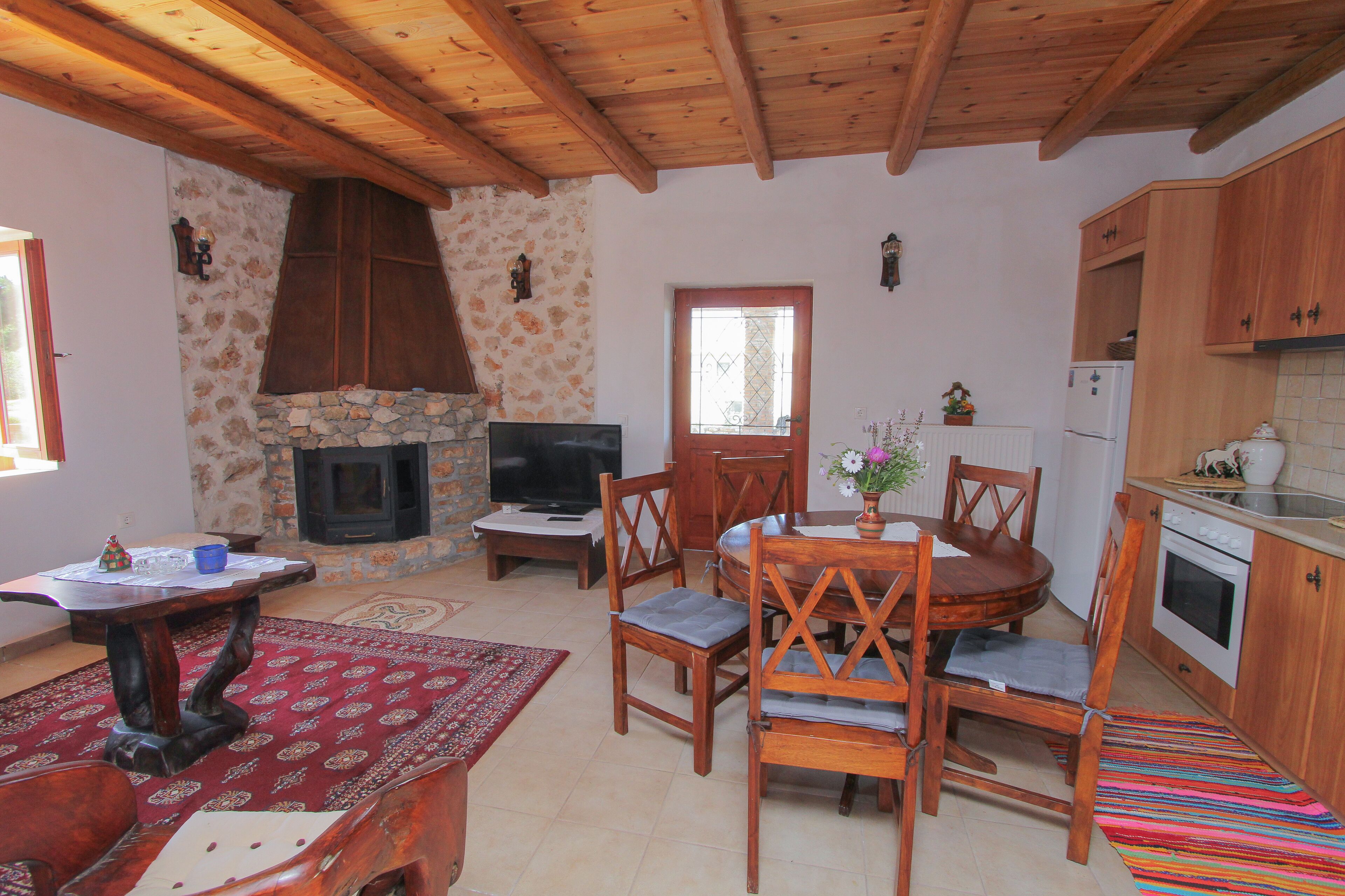 A cozy living room with a stone fireplace, wooden ceiling beams, a TV, and a dining area with a table and chairs.