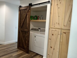 A laundry room with a wooden sliding door, a washer and dryer, and a shelf with cleaning supplies.
