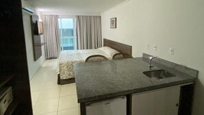 Een compacte hotelkamer met een kitchenette, een bed met hoofdeinde, een raam met gordijnen en uitzicht op de stad.