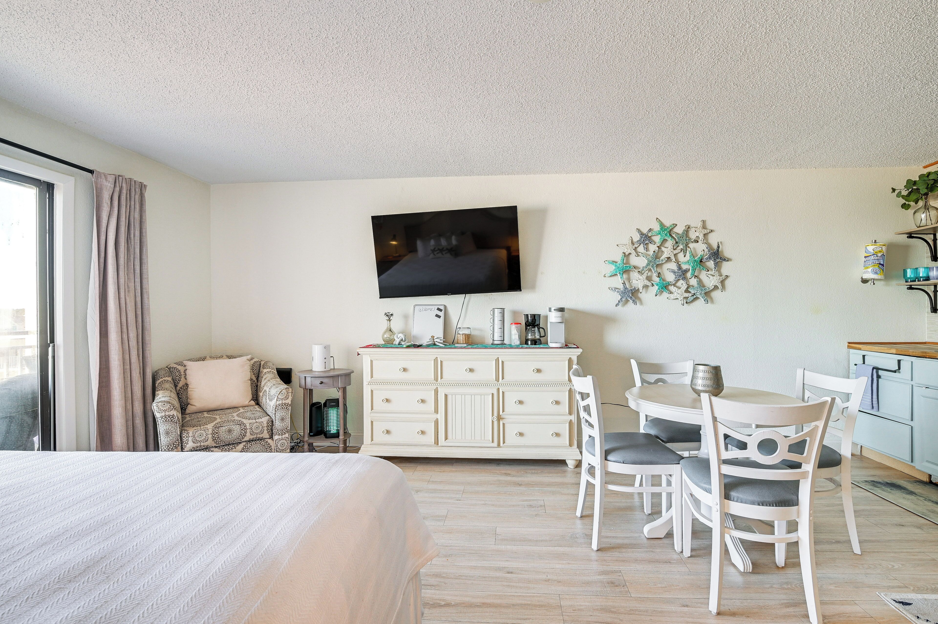 A bedroom with a bed, a TV mounted on the wall, a dresser, a chair, and a dining table with chairs.