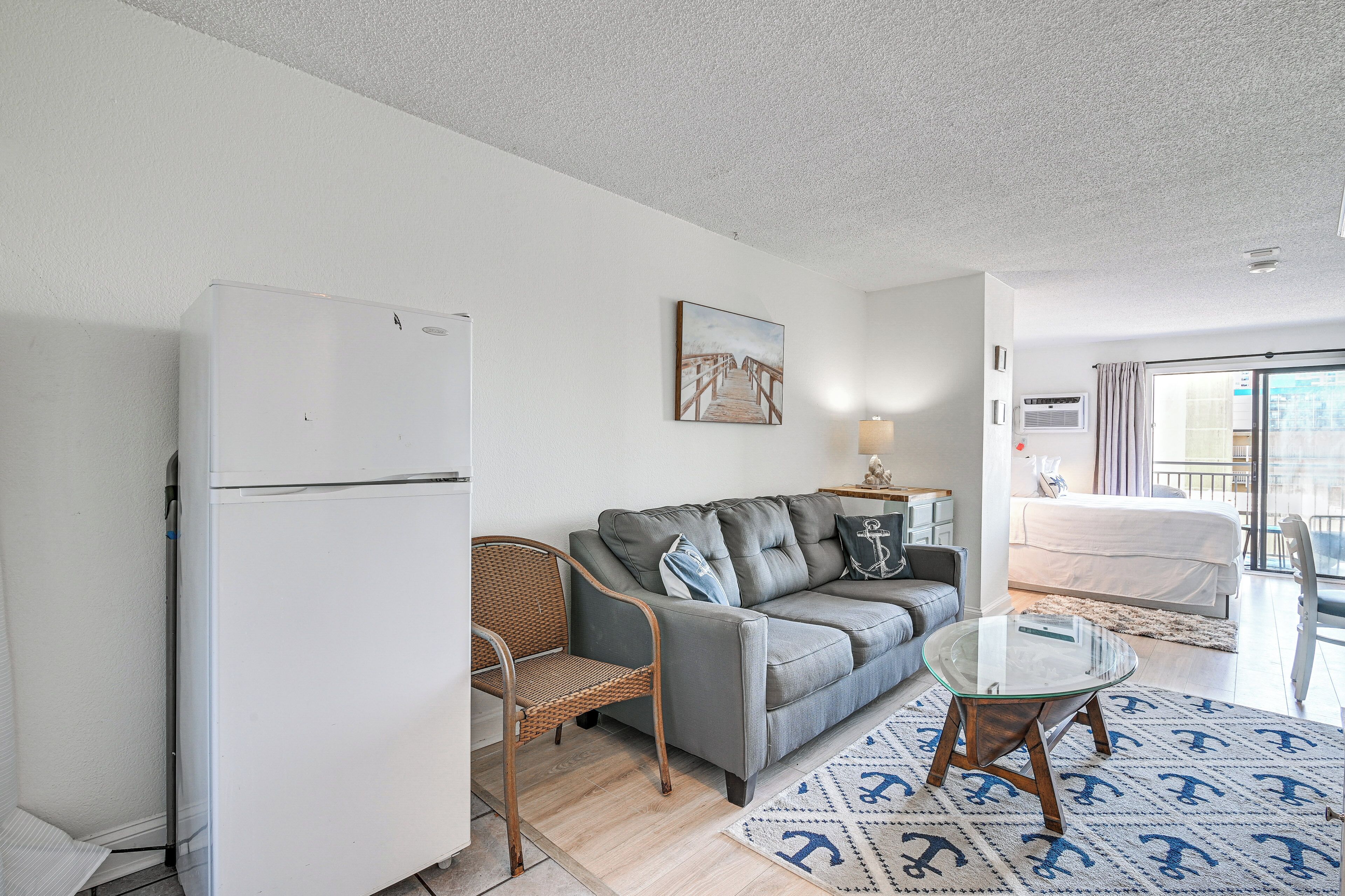 A compact living space with a grey sofa, a glass coffee table, a wicker chair, a white refrigerator, and a patterned rug.