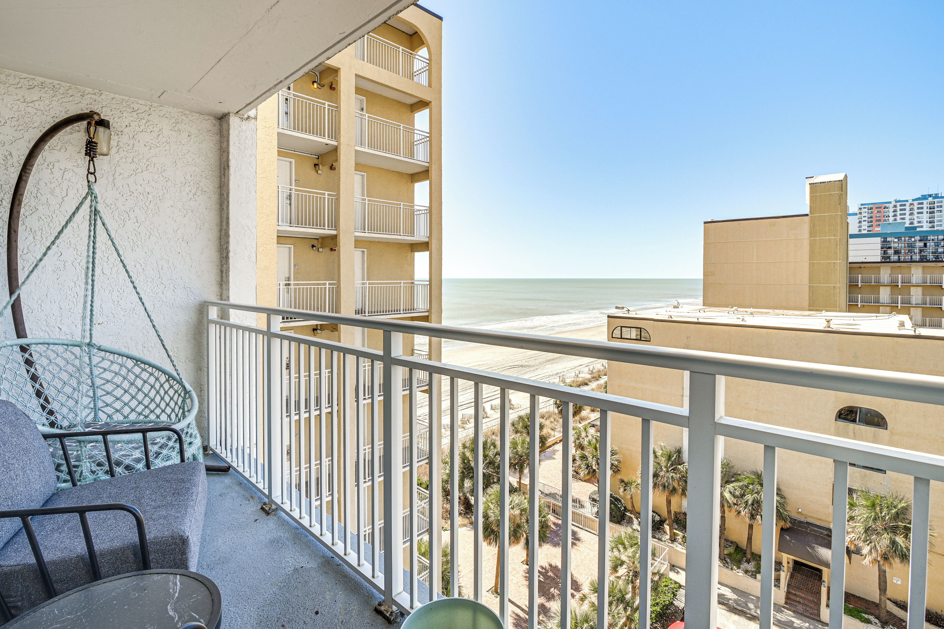 A balcony with a hanging chair and a small table, overlooking a beach and a building with multiple balconies.