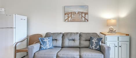 Una sala de estar con un sofá gris, una mesa de centro de vidrio, una silla de mimbre, un refrigerador blanco, un armario azul claro y un cuadro de un muelle.