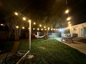 Un jardin avec un hamac, des guirlandes lumineuses, une table de pique-nique et une chaise.