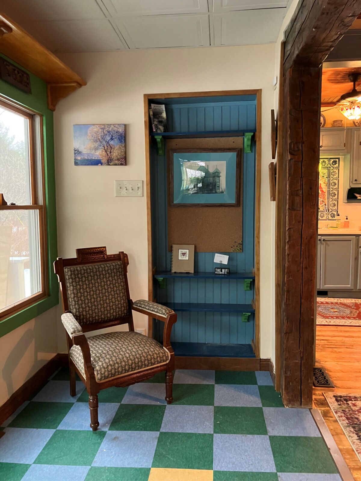 Um quarto com piso xadrez, uma cadeira de madeira, uma porta azul com espelho e uma janela com vista para árvores.