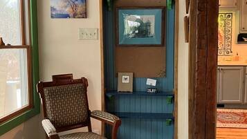 Um quarto com piso xadrez, uma cadeira de madeira, uma porta azul com espelho e uma janela com vista para árvores.