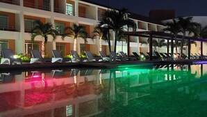 Un espace aménagé au bord de la piscine, avec des chaises longues et des palmiers, dont le reflet se projette sur les façades de l’immeuble.