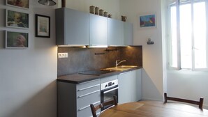 Eine kleine Küche mit grauen Schränken, einer Spüle und einem Esstisch mit Stühlen. An der Wand hängen Bilder in Rahmen und über dem Tisch hängt ein Deckenventilator.