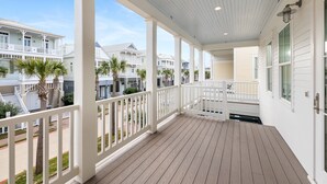 A covered porch with white railings and a wooden floor, overlooking a residential area with houses and palm trees.