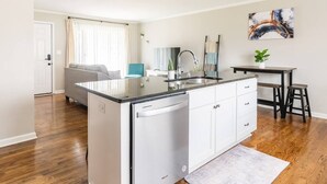 A compact kitchen with a dishwasher, a dining area with a small table and stools, and a living area with a sofa and a TV.