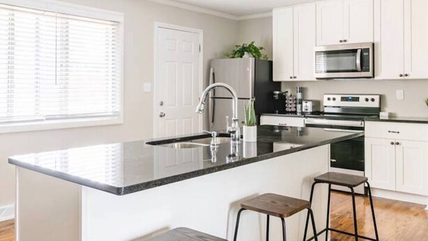 A modern kitchen with white cabinets, a black countertop, stainless steel appliances, and a window with blinds.