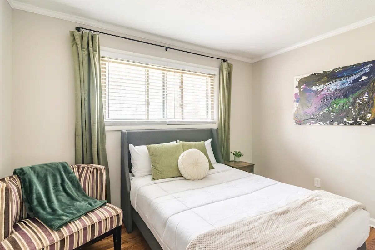 Un dormitorio con cama, una silla, ventana con cortinas y un cuadro en la pared.