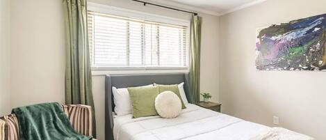 Un dormitorio con cama, una silla, ventana con cortinas y un cuadro en la pared.