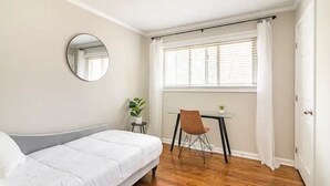 A bedroom with a bed, a round mirror, a small table with a chair, a window with blinds, and a potted plant.