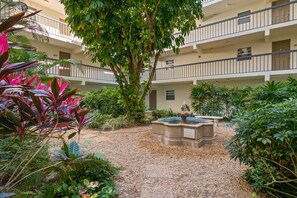 A courtyard with a fountain, surrounded by lush greenery and a multi-story building with balconies.
