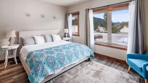 A bedroom with a bed, a nightstand, a blue chair, a window with curtains, and a view of the outside.
