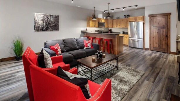 A modern living room with a black sofa, red chairs, a coffee table, and a kitchen area with wooden cabinets and stainless steel appliances.