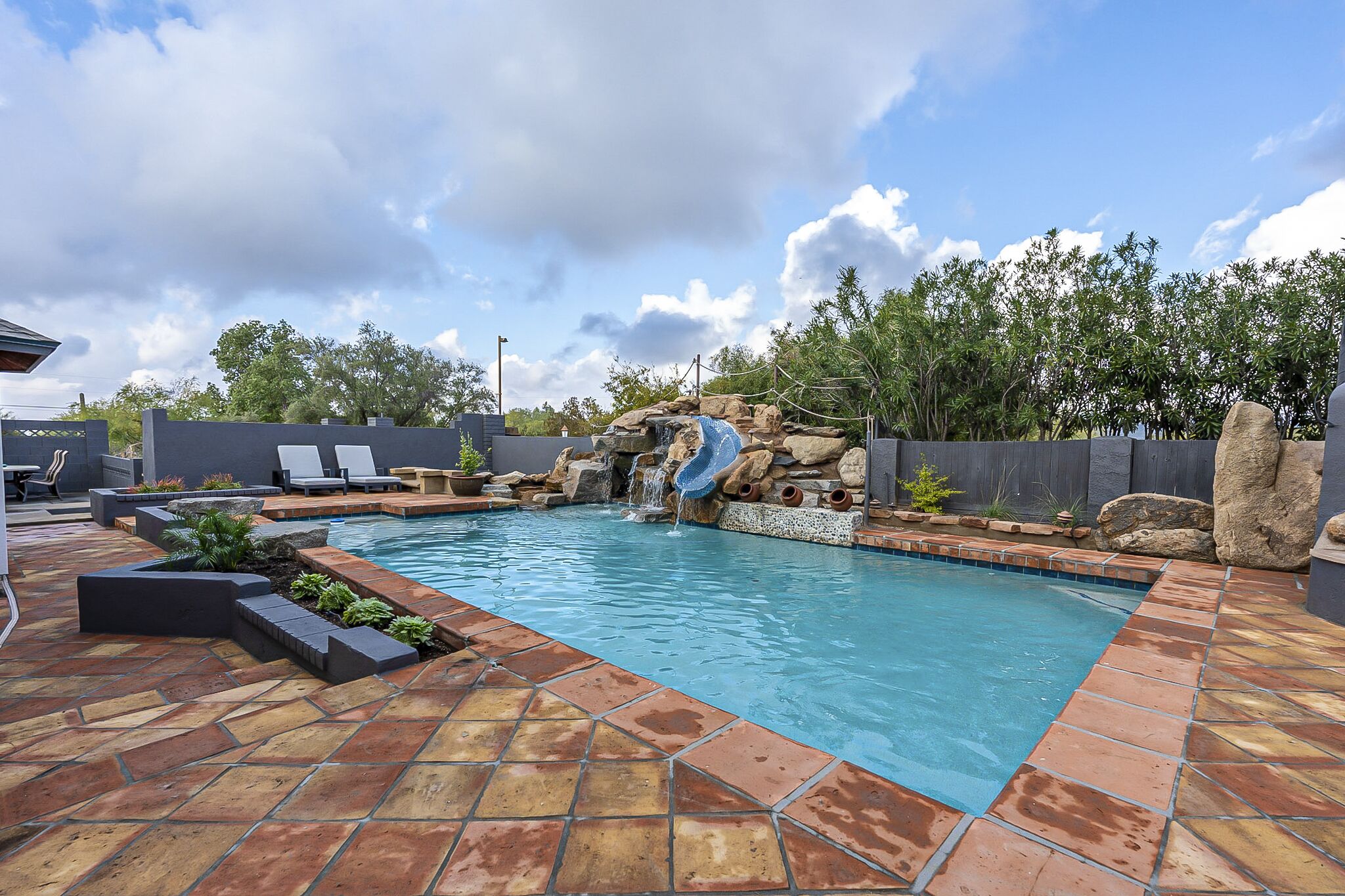 Ein Pool mit Wasserfall, umgeben von einer steinbelagten Terrasse und einem Holenzaun.