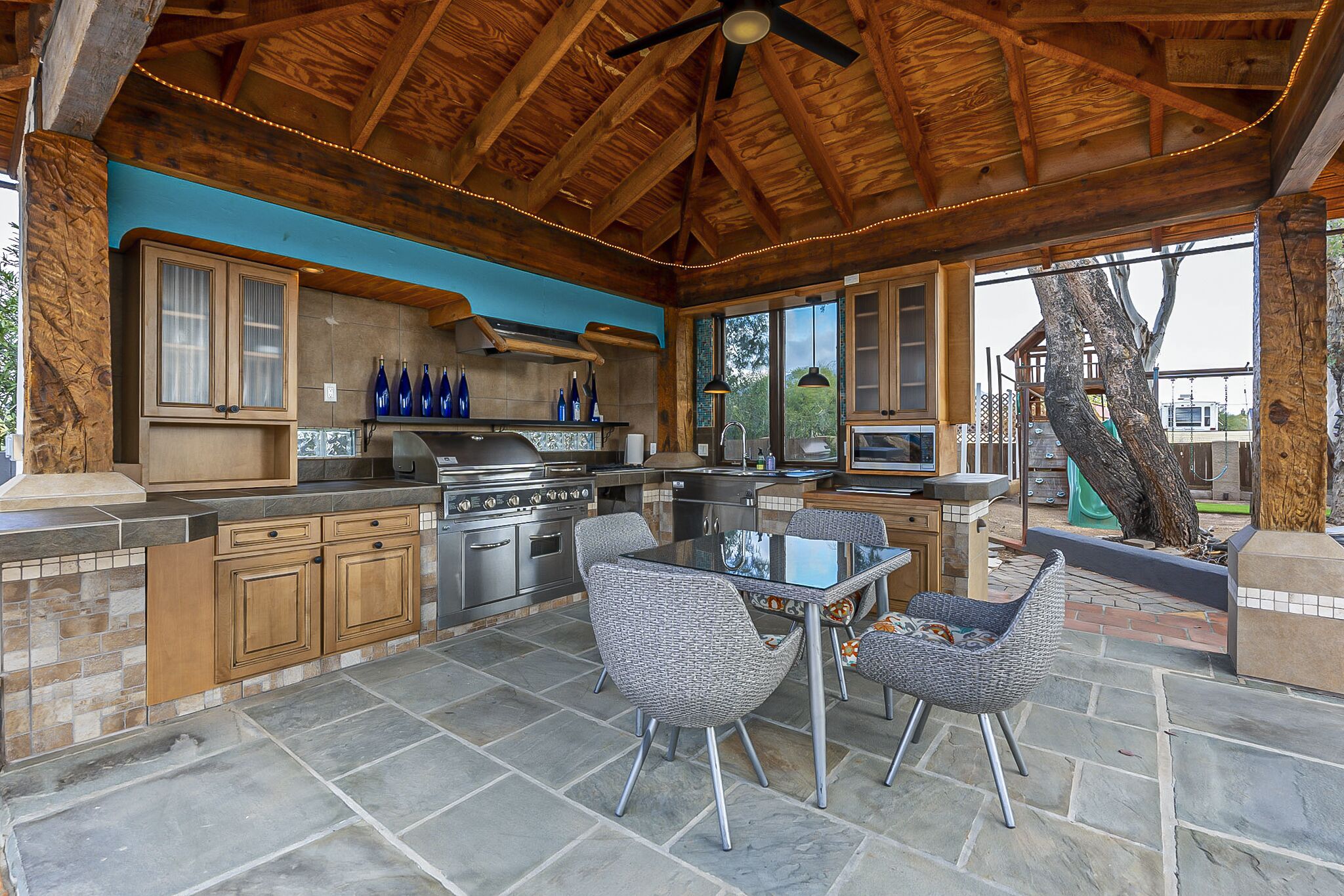 A covered outdoor kitchen with stone flooring, wooden cabinets, and a dining area with chairs.