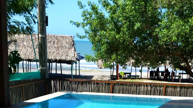 Una piscina azul cristalina con una terraza de madera y una estructura de techo de paja al fondo.