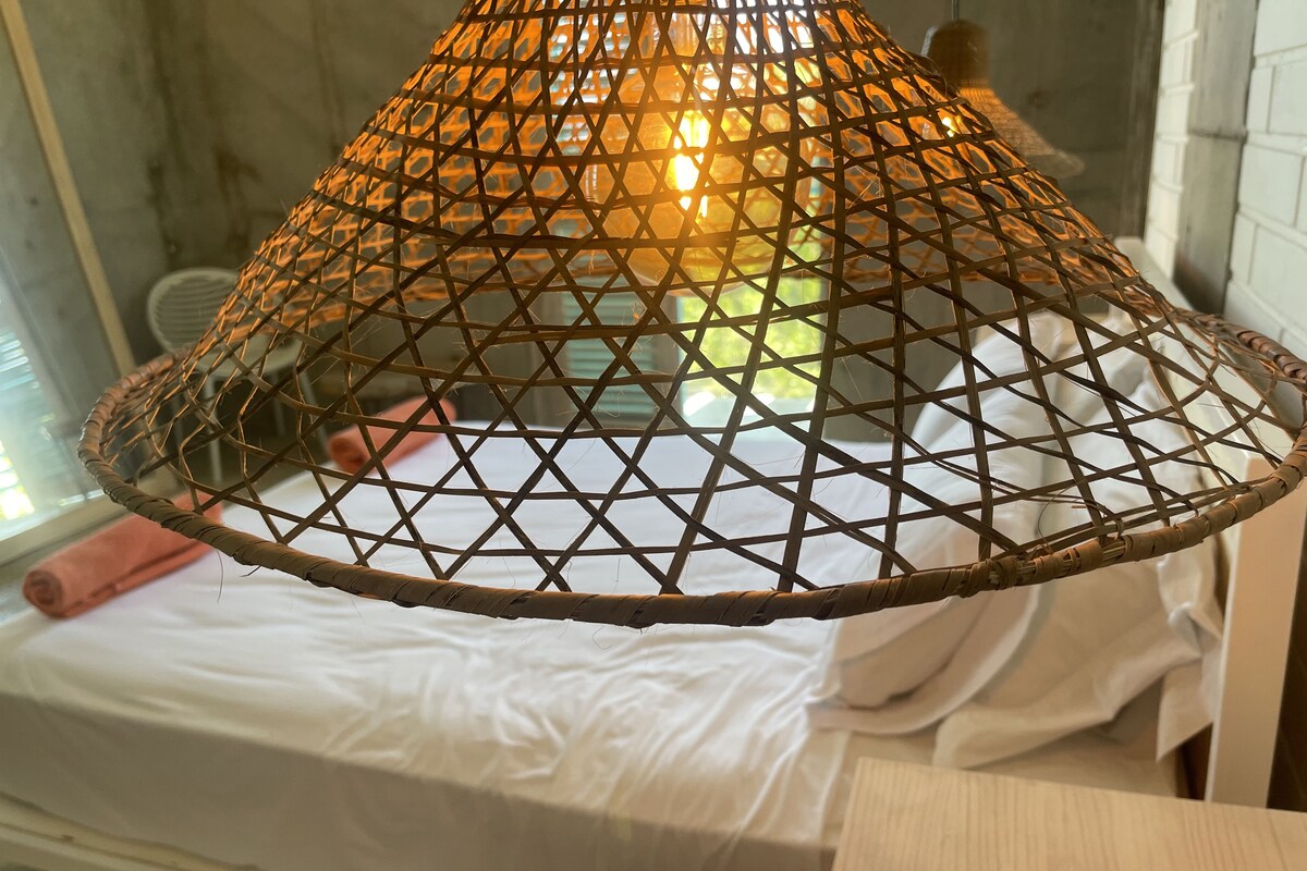 Une pièce comprenant un lit, une table de chevet en bois et un luminaire suspendu en fibres tressées.