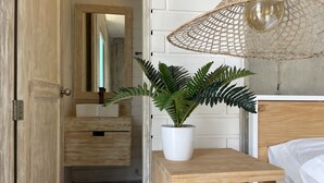 Un baño con tocador de madera, un lavamanos blanco, un espejo, una planta en maceta y una lámpara colgante de ratán.