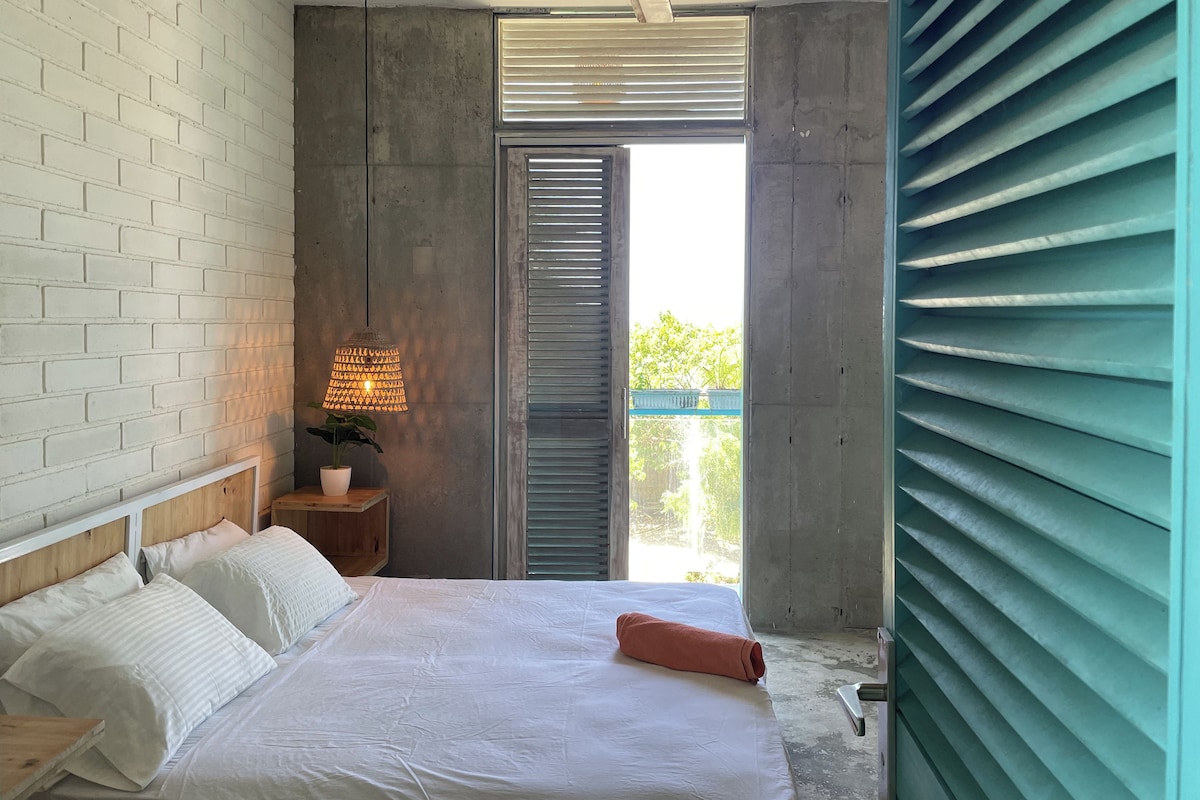 Une chambre à coucher avec un lit, des oreillers blancs, une table de chevet avec une lampe et une fenêtre avec des stores.