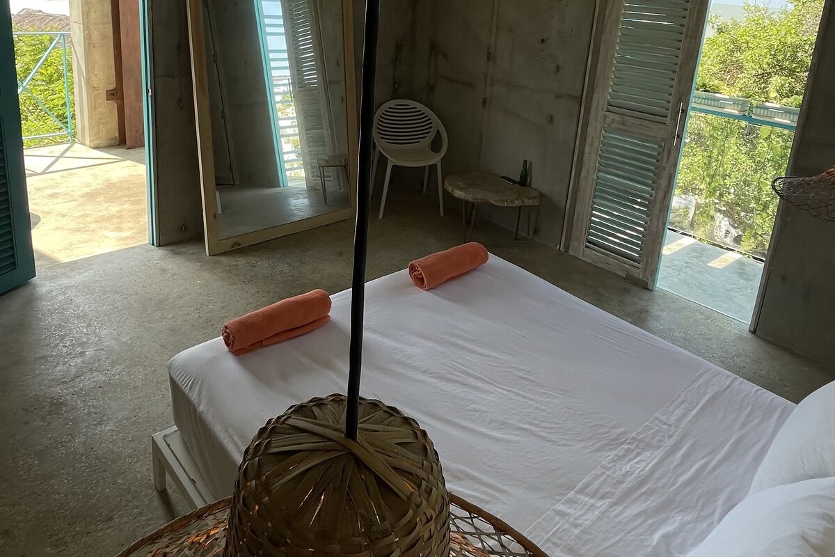 A bedroom with a bed, a woven basket, a chair, and a view of greenery outside.