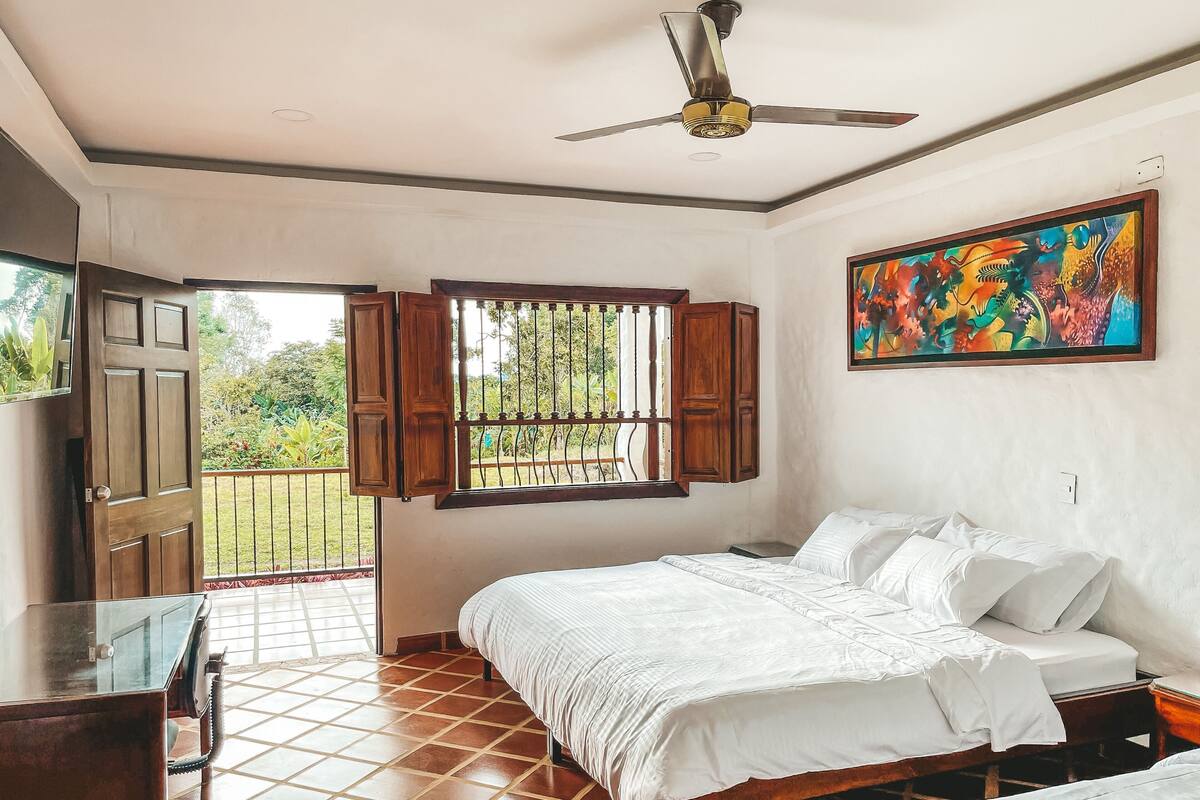 Une chambre à coucher avec un lit, un ventilateur de plafond, une fenêtre donnant sur un beau paysage et un tableau accroché au mur.