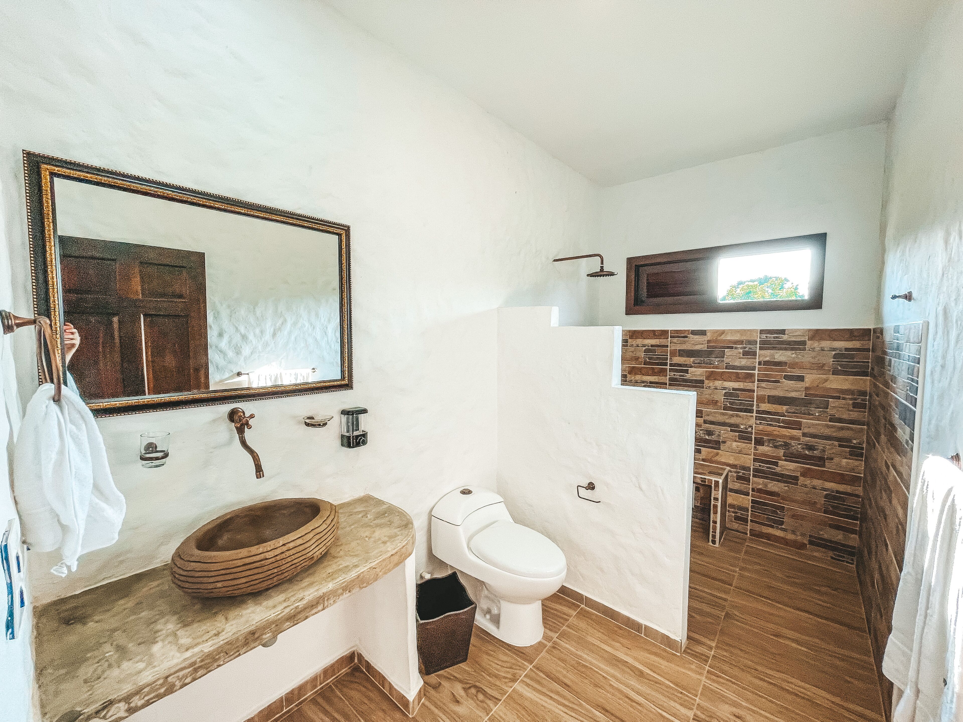 Un baño con lavamanos de mármol, un cuenco de madera, inodoro blanco, ducha con pared de piedra y un espejo grande con marco de madera.