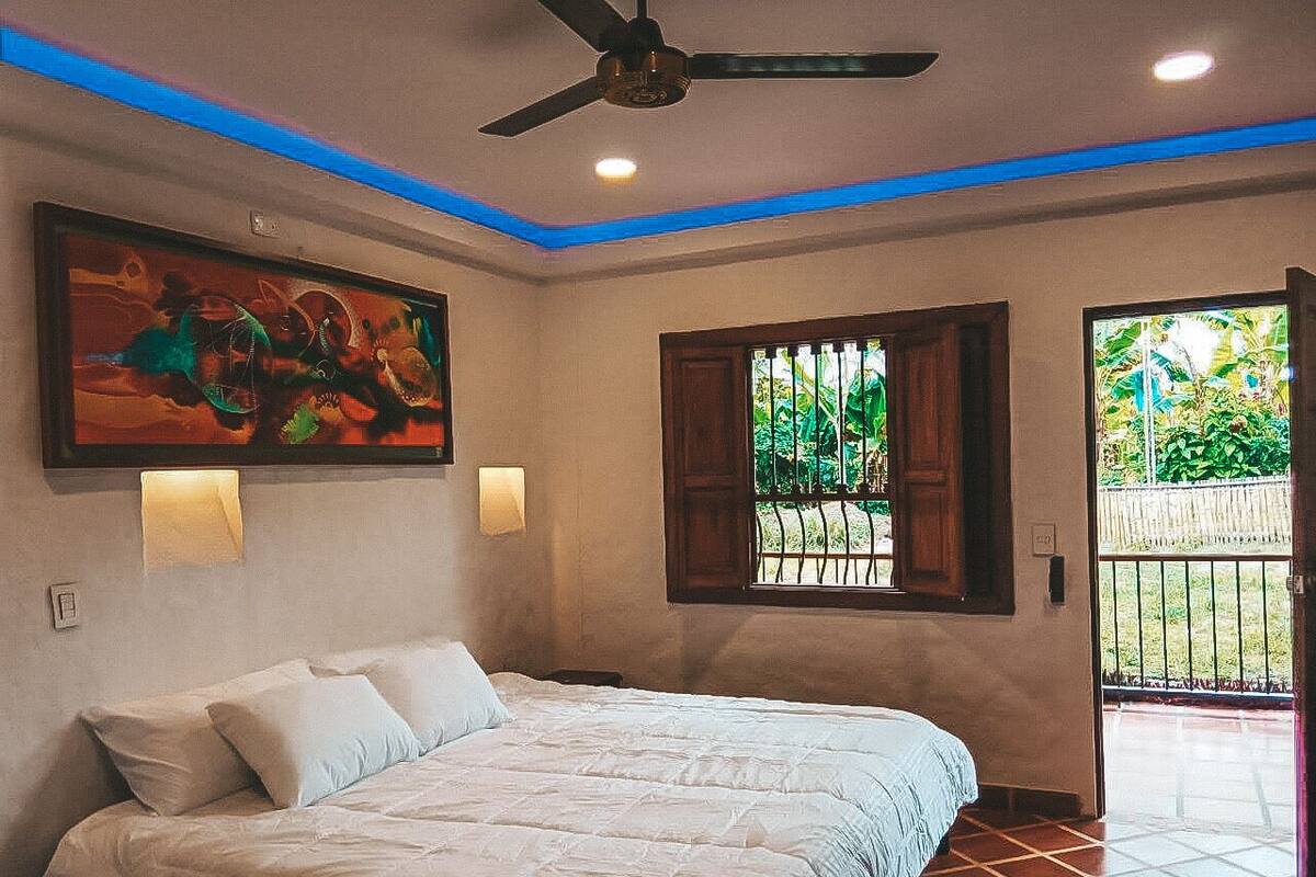 Une chambre à coucher avec un lit, un ventilateur de plafond, un tableau et une fenêtre donnant sur un paysage verdoyant.