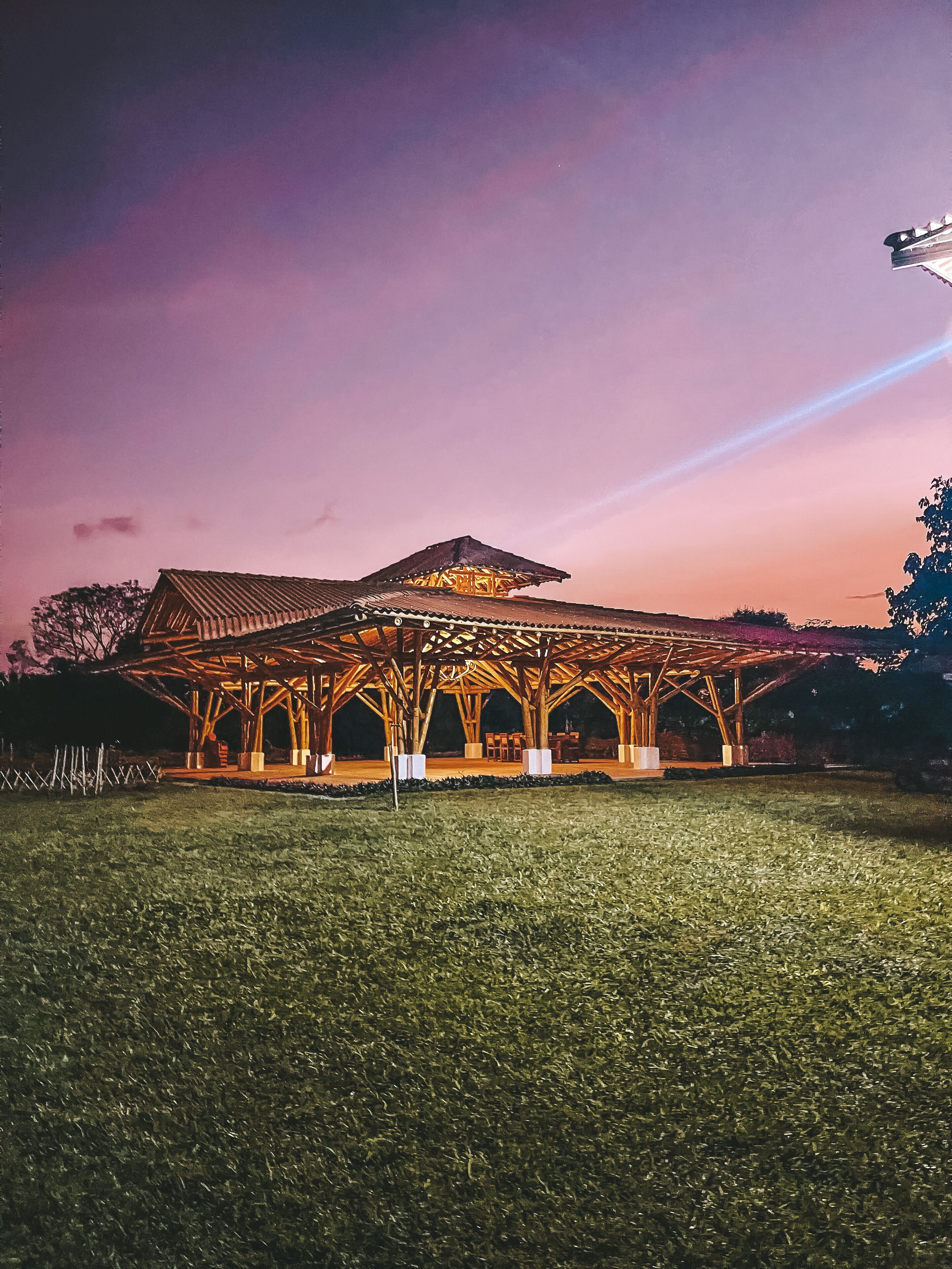 A large, open pavilion with a wooden roof structure and multiple seating areas, set in a grassy field.