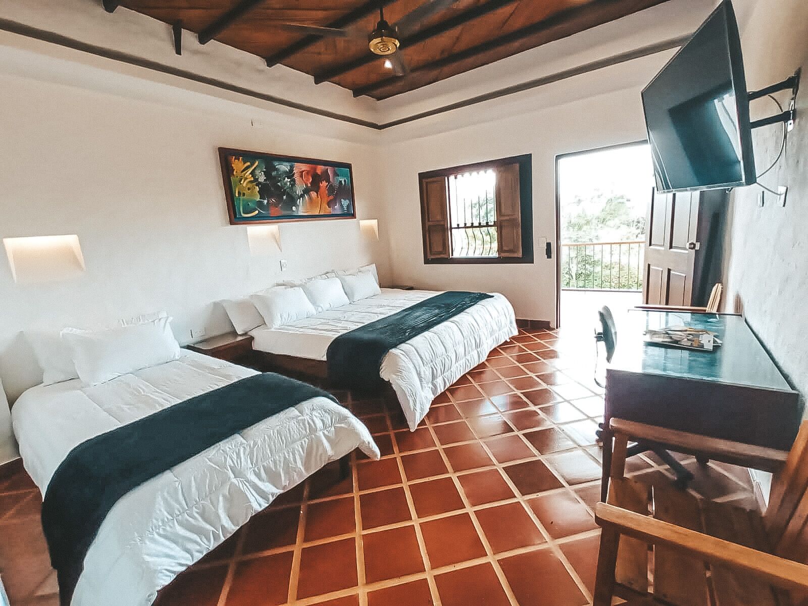 A hotel room with two beds, a desk, a TV, a painting, and a balcony.