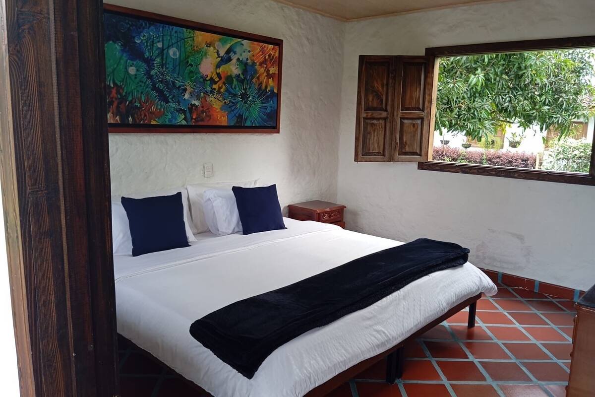 Une chambre avec un grand lit, une tête de lit en bois, un tableau au-dessus du lit, une fenêtre donnant sur un paysage verdoyant et un sol carrelé.