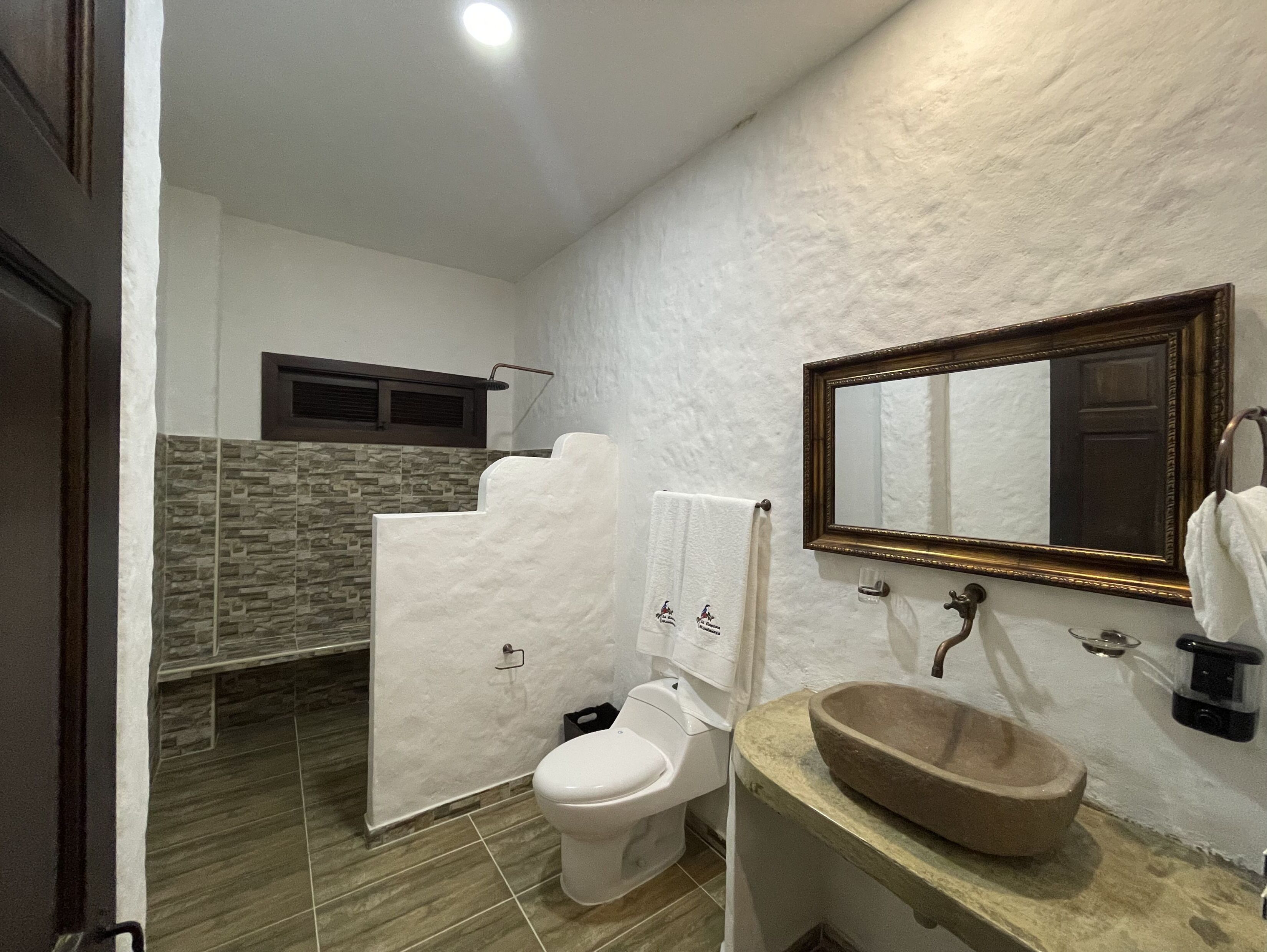 A bathroom with a stone tile floor, a white toilet, a beige sink, and a mirror with a gold frame.