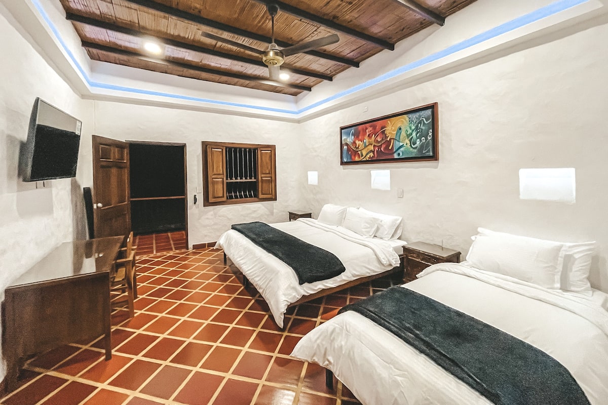 Une chambre d’hôtel avec deux lits, une télévision, un ventilateur de plafond et un tableau au mur.