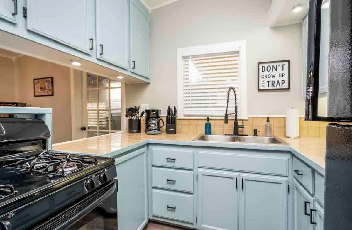 Una cocina con armarios azul claro, estufa negra, lavaplatos y ventana con persianas.