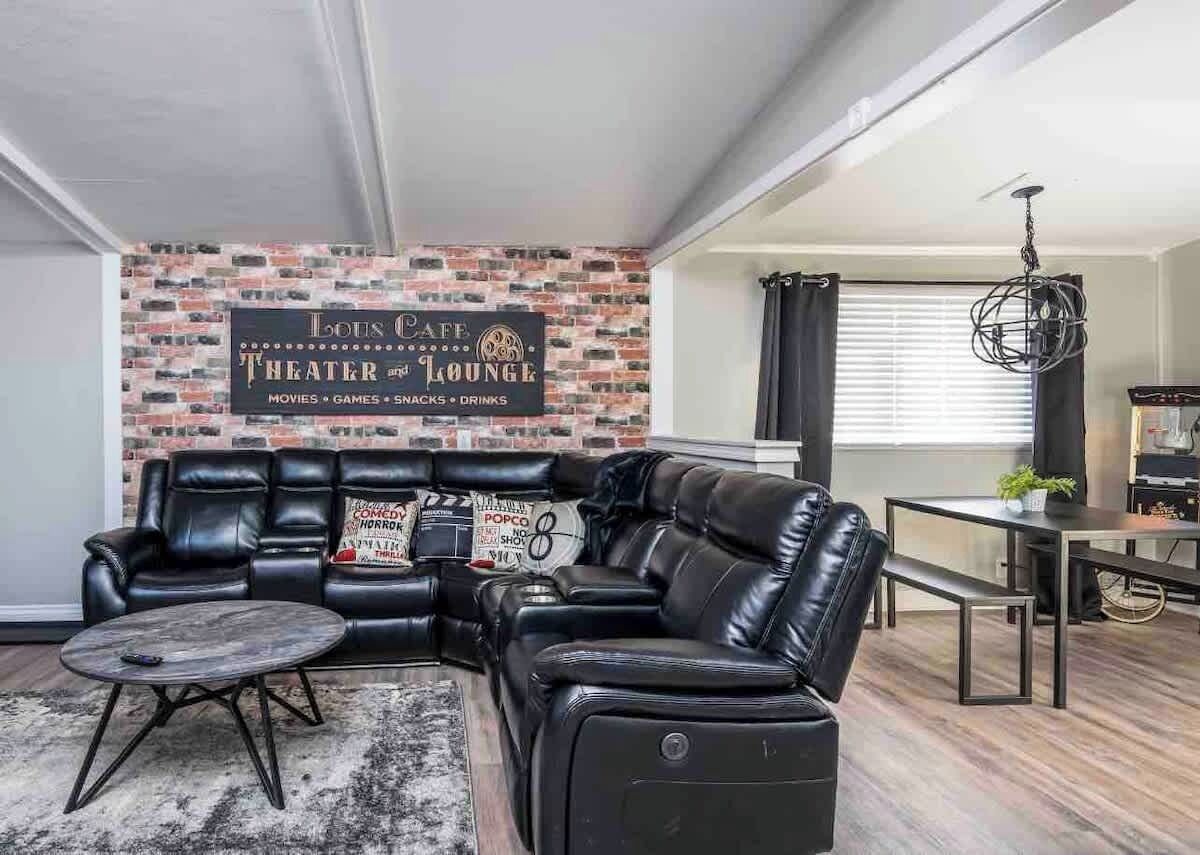 A living room with a black leather sectional sofa, a coffee table, a brick-patterned wall, and a sign for 'Louis Cafe Theater and Lounge' offering movies, games, snacks, and drinks.