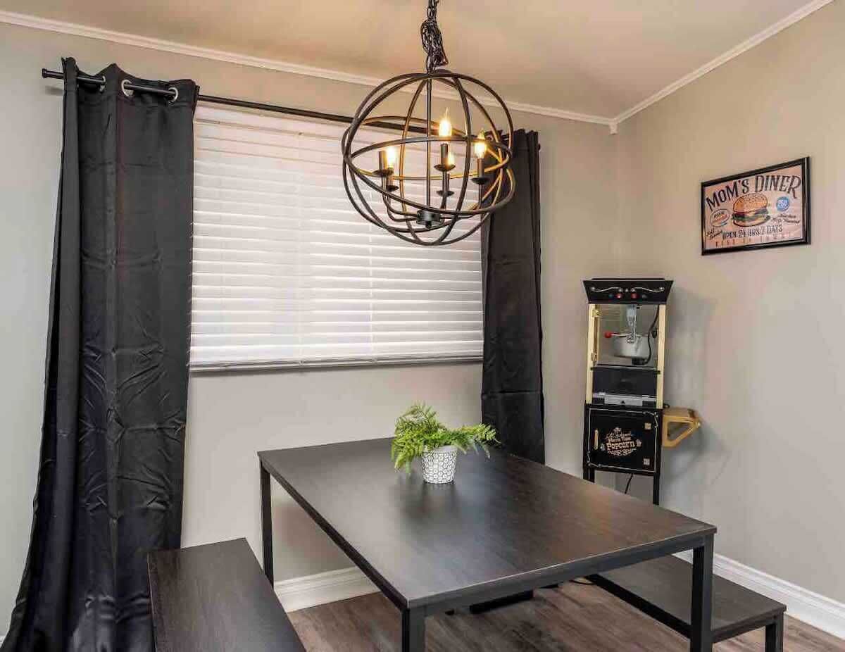 A dining area with a table, black curtains, a hanging light fixture, a small plant, and a popcorn machine.