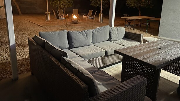 Un área de patio al aire libre con un sofá, sillas y una mesa. Se ven luces colgantes y un hogar para fuego al fondo.