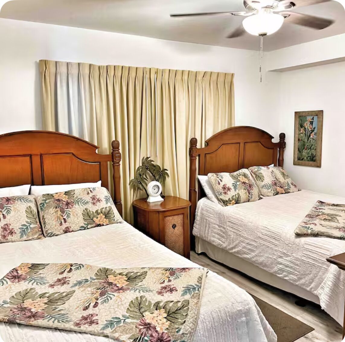 Ein Hotelzimmer mit zwei Holzbetten, beigen Vorhängen, einem Deckenventilator, einem kleinen Nachttisch mit einer Pflanze und einem an der Wand hängenden Gemälde.