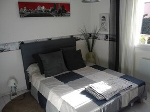 Une chambre à coucher comprenant un lit, une table de chevet, une lampe, un vase avec des plantes et une fenêtre donnant sur l’extérieur.