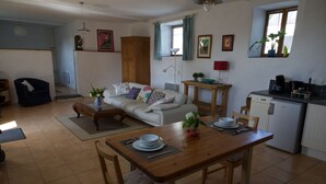 A dining area with a wooden table set for two, a sofa with colorful cushions, a kitchen area with a refrigerator, and a window with curtains. - Idyllic Country Location for couples, central, peaceful, 5 mins from shops, WIFI (LOPEREC)
