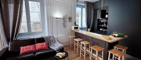 A modern living room with a dark-colored sofa, a wooden coffee table, and a kitchen area with a bar counter and stools.