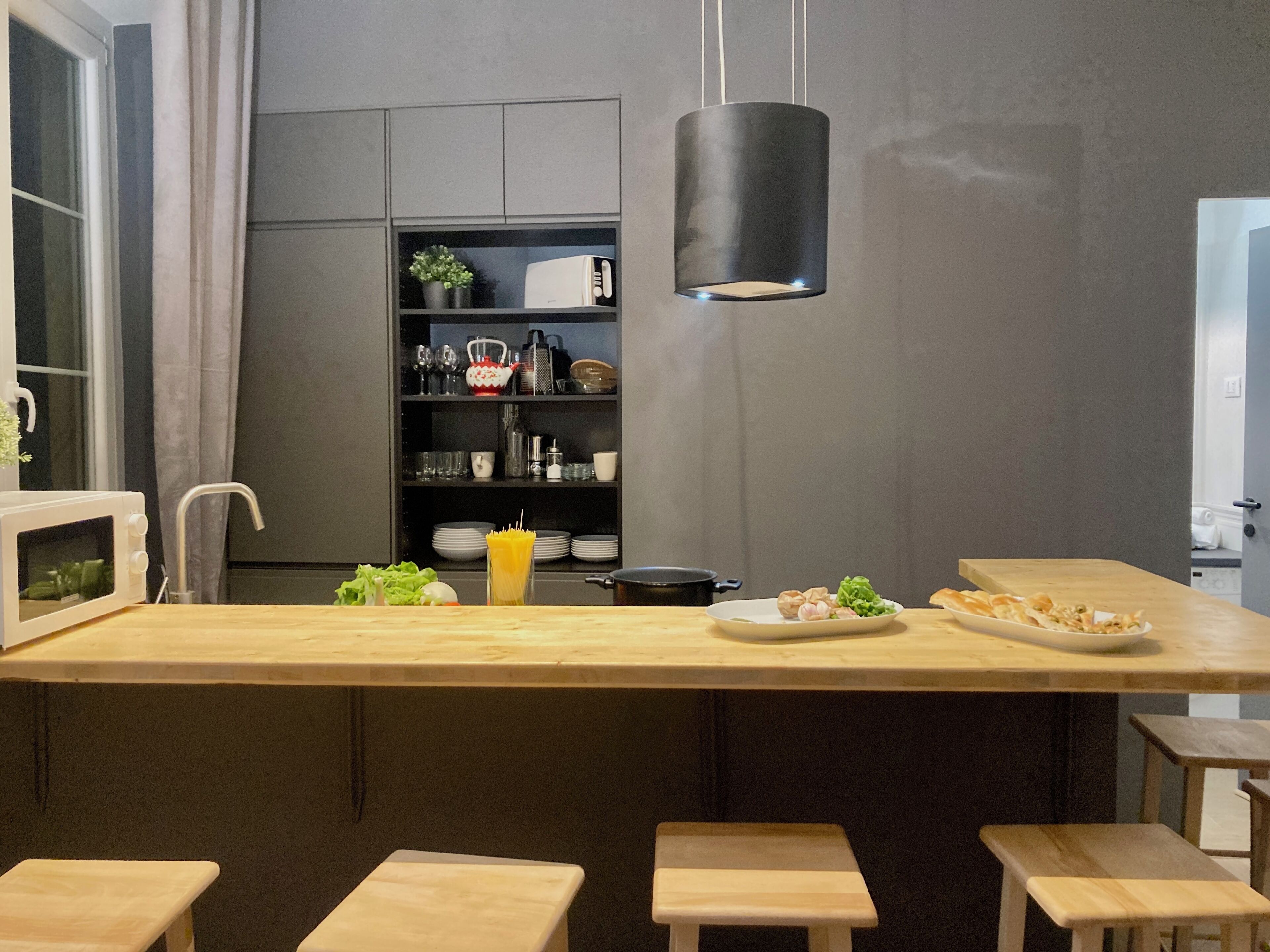 A modern kitchen with a central island, wooden stools, and a hanging light fixture.