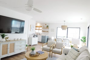 A modern living room with a flat-screen TV, a dining area with chairs, a kitchen in the background, and a ceiling fan.