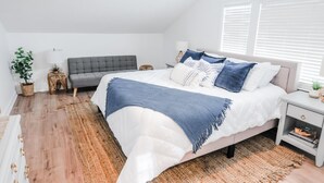 Une chambre avec un grand lit, un canapé gris, une petite table, une lampe et une plante.
