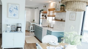 Une cuisine moderne avec des meubles blancs, un coin repas doté d’une table en bois et de chaises blanches, et un lustre suspendu en osier.