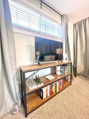 A living room with a TV on a wooden stand, a bookshelf with books, and a window with blinds.